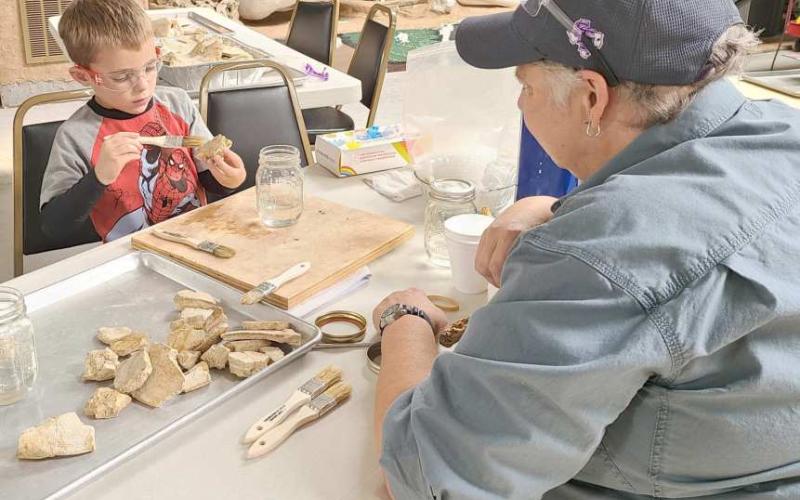 FOSSIL PRESERVATION—During Archeology Day earlier this month at the Museum of the Plains, kids and parents got to learn more about archeology and participate in activities like ledger art, painted teepees, spear throwing with an atlatl spear, clay pottery, and an archeology dig. Pictured are museum director Stacey Brown supervising Jax Hinshaw, age 5, as he learns about preserving fossils with resin.
