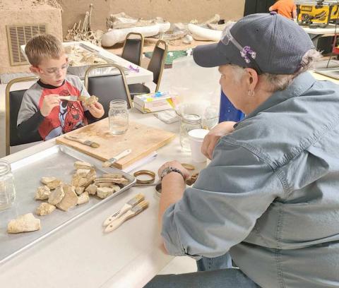 FOSSIL PRESERVATION—During Archeology Day earlier this month at the Museum of the Plains, kids and parents got to learn more about archeology and participate in activities like ledger art, painted teepees, spear throwing with an atlatl spear, clay pottery, and an archeology dig. Pictured are museum director Stacey Brown supervising Jax Hinshaw, age 5, as he learns about preserving fossils with resin.