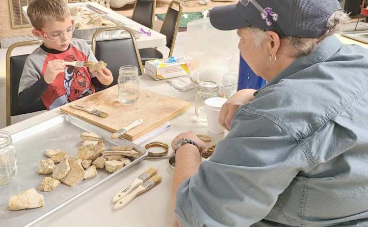 FOSSIL PRESERVATION—During Archeology Day earlier this month at the Museum of the Plains, kids and parents got to learn more about archeology and participate in activities like ledger art, painted teepees, spear throwing with an atlatl spear, clay pottery, and an archeology dig. Pictured are museum director Stacey Brown supervising Jax Hinshaw, age 5, as he learns about preserving fossils with resin.